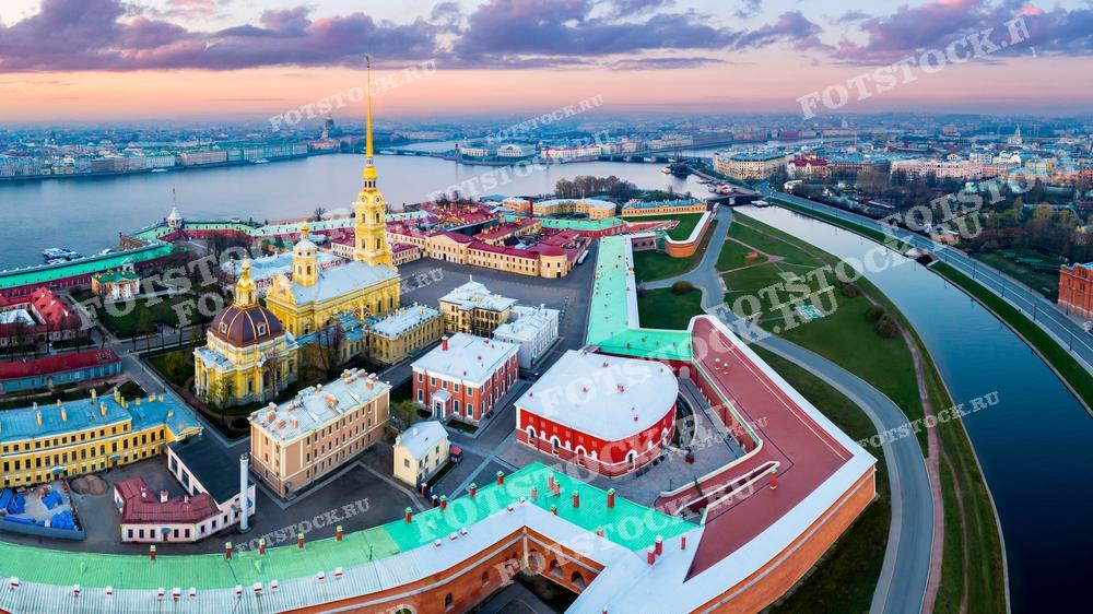 На этой фотографии мы видим величественный Петропавловский собор и крепость в Санкт-Петербурге. Золотой шпиль собора возвышается над окружающими зданиями, а река Нева извивается вдоль крепостных стен, создавая красивый контраст между водной гладью и ярко-зелёными лужайками. 
Фотография подойдёт для исторических публикаций, туристических материалов, постеров, а также для использования в образовательных целях, чтобы показать уникальность русской архитектуры и планировки городов.